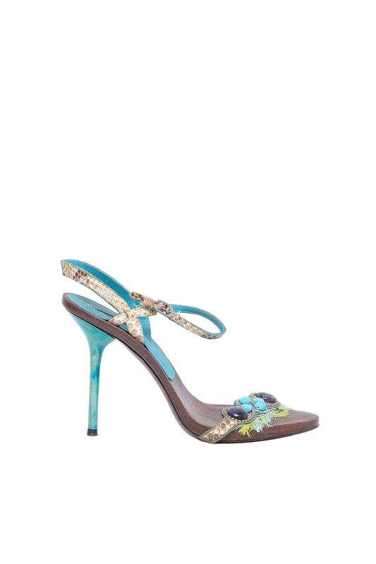 A single women's high-heeled sandal with a brown leather base, turquoise blue straps, and a decorative floral embellishment on the toe strap, shown against a light blue background.